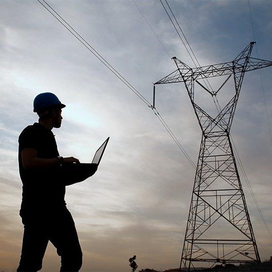 Network engineer working in the field, wearing a hard hat and holding a laptop, looking at a network tower at dusk 